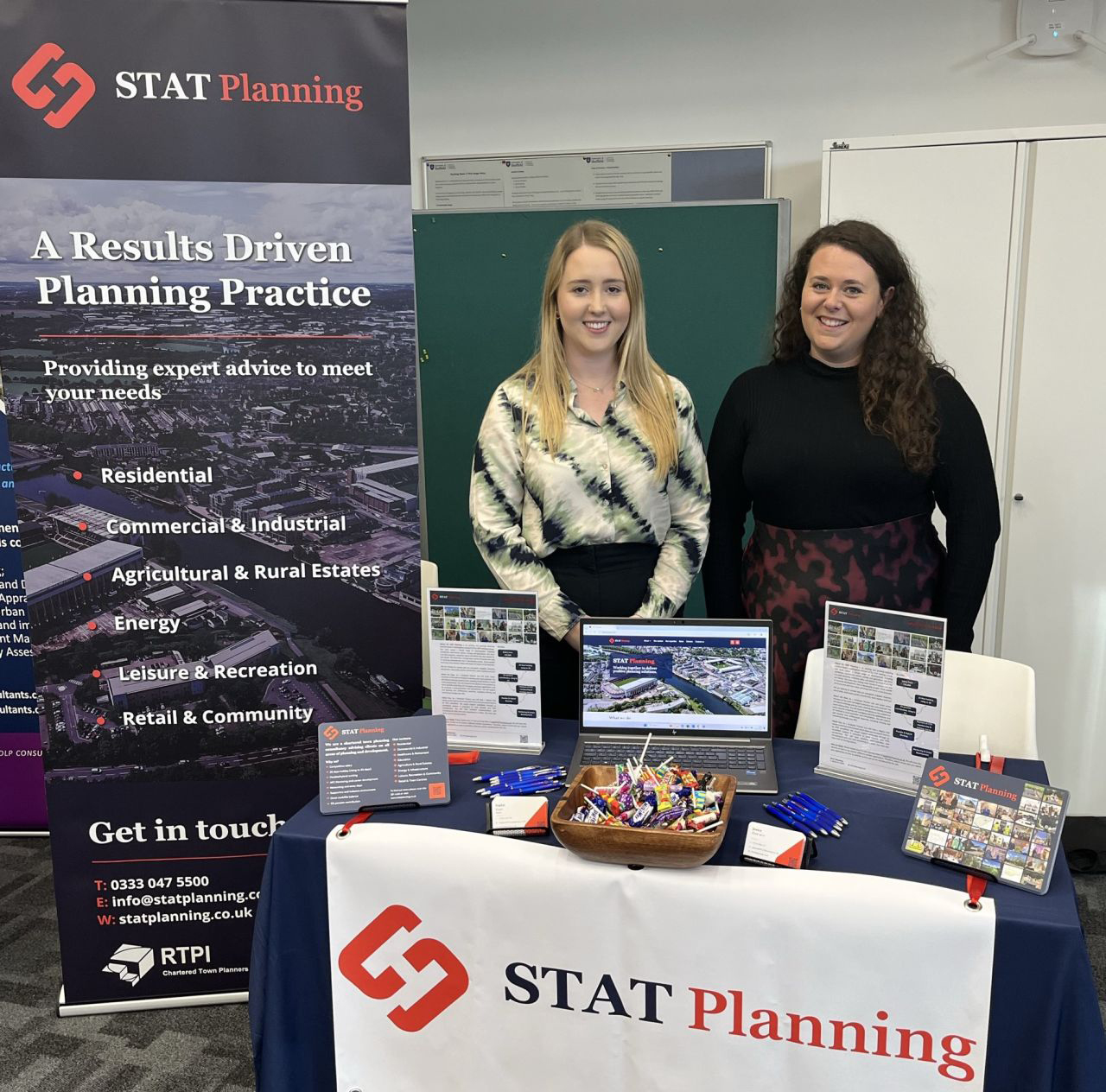 Two professionals from STAT Planning standing at an exhibition booth with a banner and laptop, showcasing planning consultancy services.