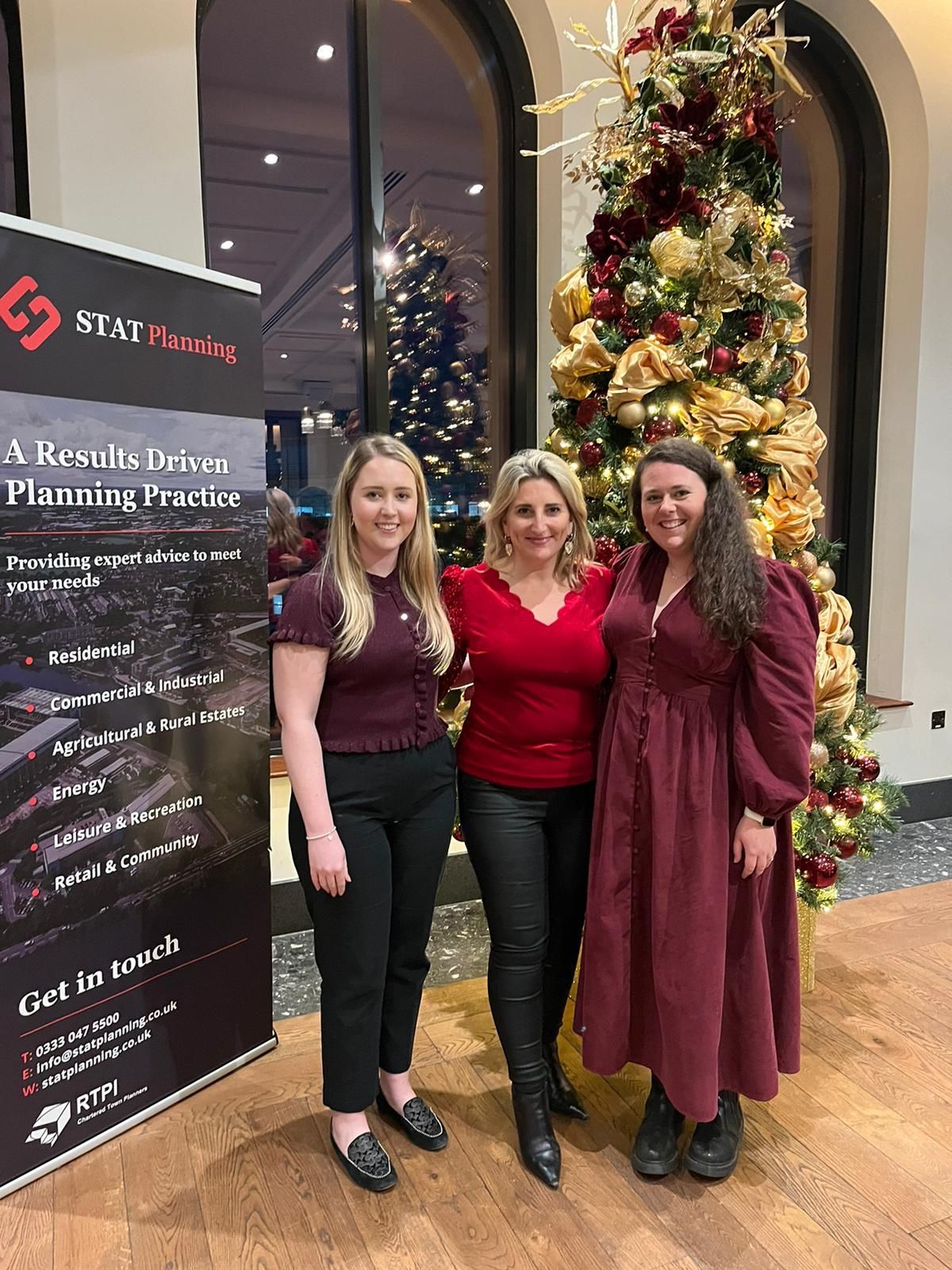 Three professionals from STAT Planning standing in front of a banner and a decorated Christmas tree, showcasing planning consultancy services during a holiday event.
